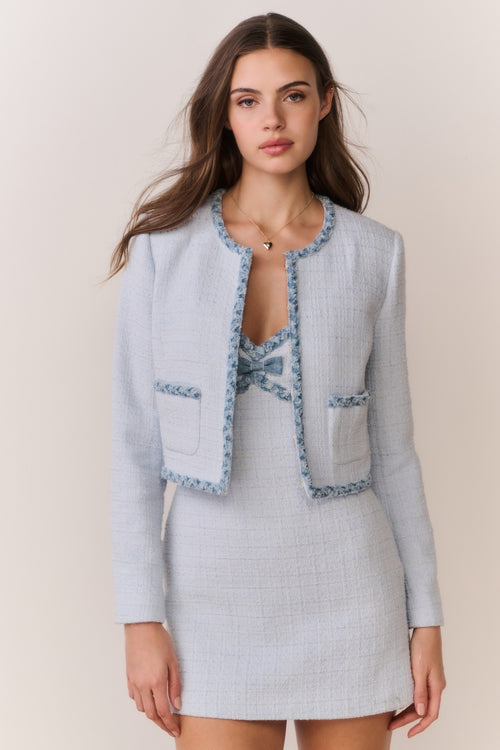 Woman wearing a light blue tweed jacket with decorative trim and matching mini dress on a plain background