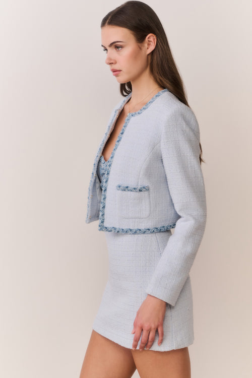 Woman wearing a light blue tweed jacket with decorative trim and matching mini dress on a plain background