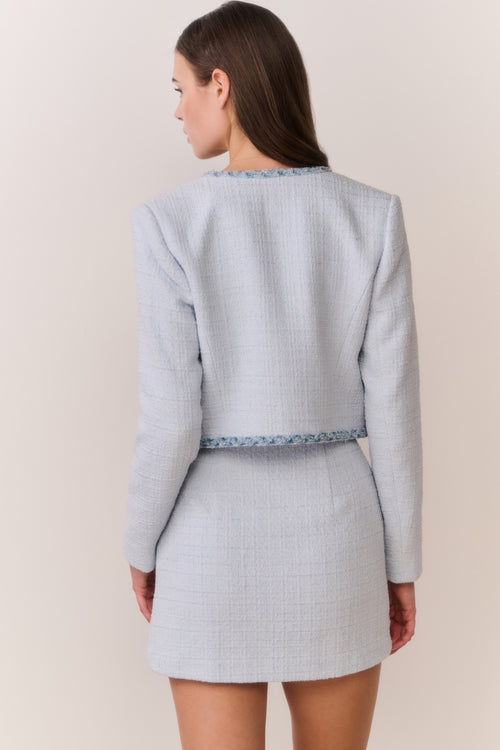 Woman wearing a light blue tweed jacket with decorative trim and matching mini dress on a plain background