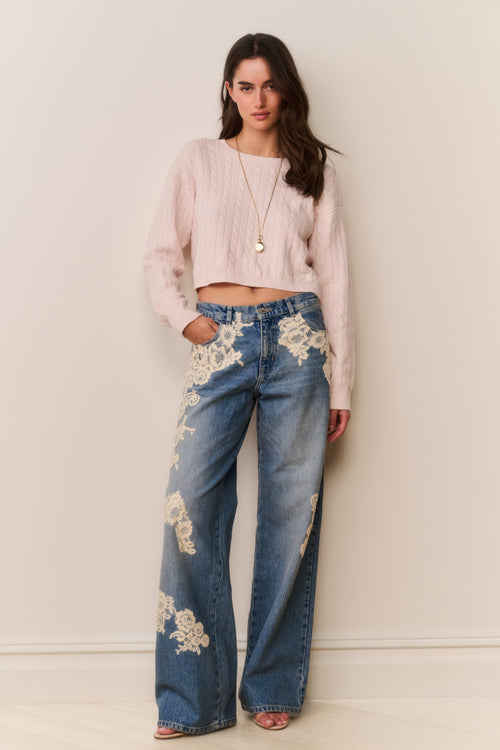 Woman wearing light pink sweater and blue jeans with floral lace appliqué against a beige background