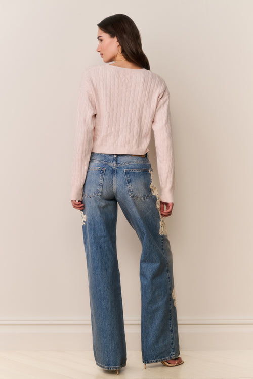 Woman wearing light pink sweater and blue jeans with floral lace appliqué against a beige background