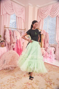 Young girl in a black top and light green skirt standing in a room with pink curtains and decor.