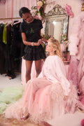 Woman in a black outfit standing next to another woman in a pink dress with feathers, in a room with floral decorations and a mirror.