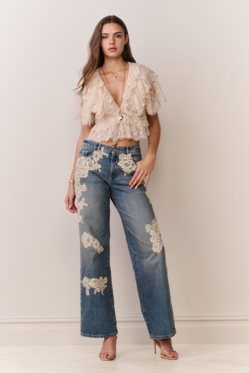 Woman wearing a lace top and jeans with floral embroidery against a plain background