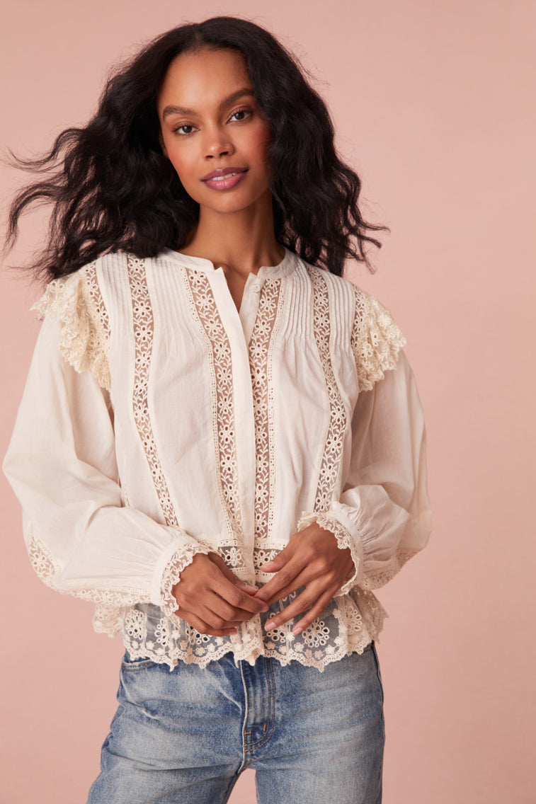 White long sleeve blouse featuring mesh and laces throughout. Has delicate mesh ruffles at the shoulder of the long blouson sleeves, shirred mesh ruffles with scallop edges at the hem, and a hidden placket with buttons down center front.