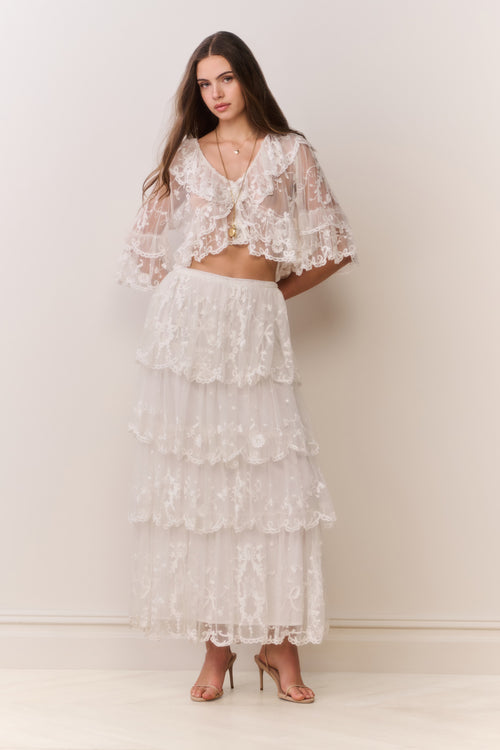 Woman wearing a white lace top and matching tiered skirt against a plain background
