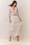 Woman wearing a white lace top and matching tiered skirt against a plain background
