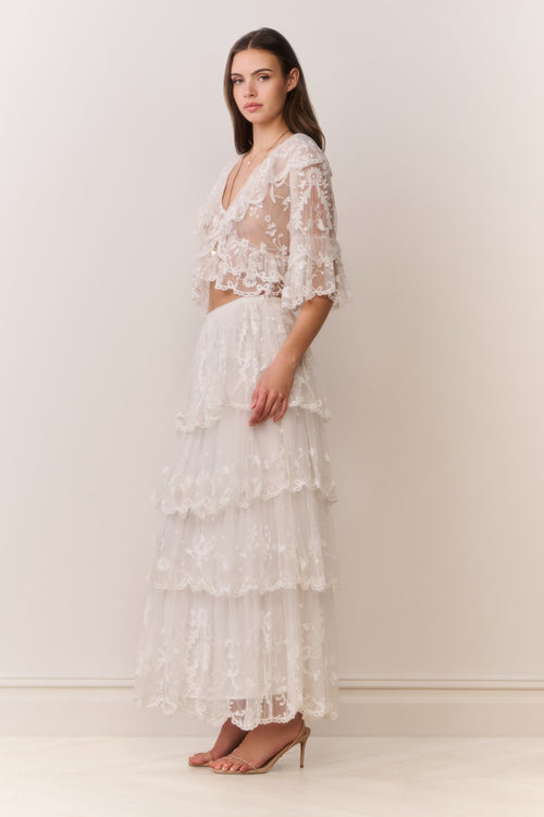 Woman wearing a white lace top and matching tiered skirt against a plain background