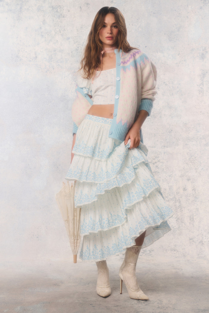 Woman wearing a layered outfit with a white top, light blue cardigan, and ruffled skirt against a neutral background.