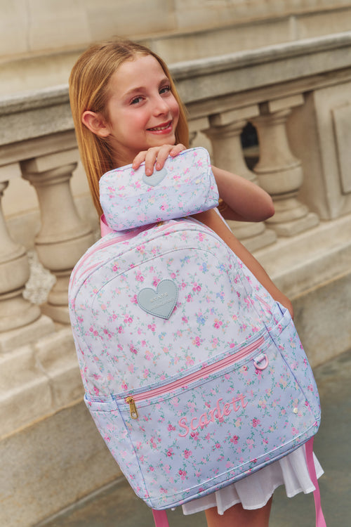Girl holding a light blue backpack with pink floral pattern and gray heart design.
