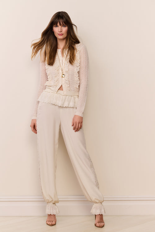 Woman wearing a cream sweater and cream pants with ruffle cuff against a plain background