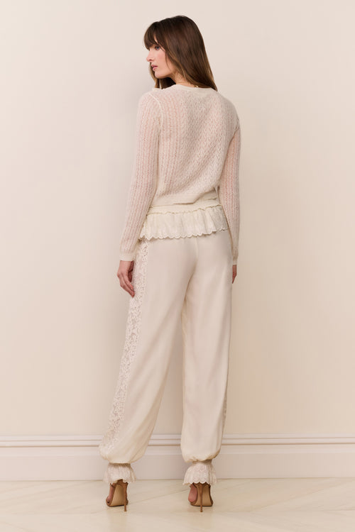Woman wearing a cream sweater and cream pants with ruffle cuff against a plain background
