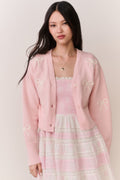 Woman wearing a pink cardigan with bow details and a pink and white lace trim skirt on a plain background