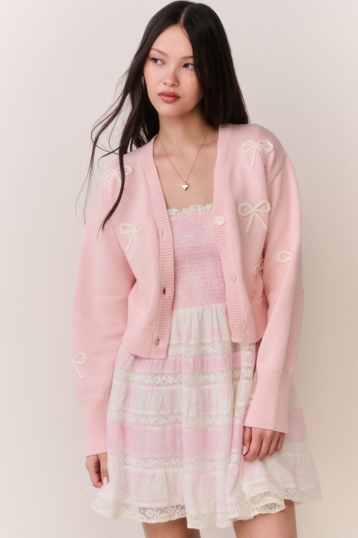 Woman wearing a pink cardigan with bow details and a pink and white lace trim skirt on a plain background