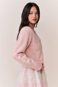 Woman wearing a pink cardigan with bow details and a pink and white lace trim skirt on a plain background