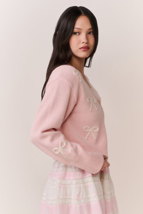 Woman wearing a pink cardigan with bow details and a pink and white lace trim skirt on a plain background