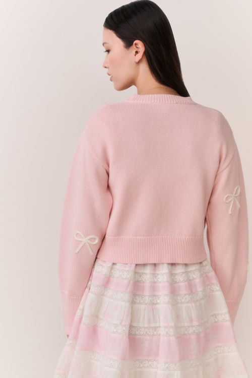 Woman wearing a pink cardigan with bow details and a pink and white lace trim skirt on a plain background