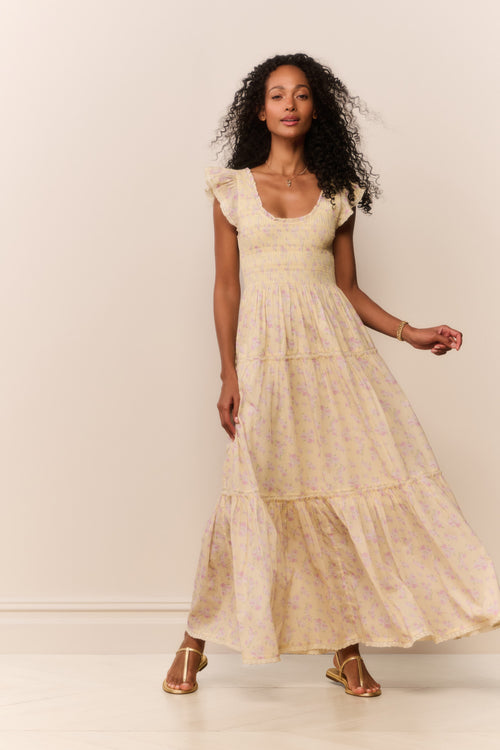 Woman wearing a long, yellow dress with floral patterns against a plain background