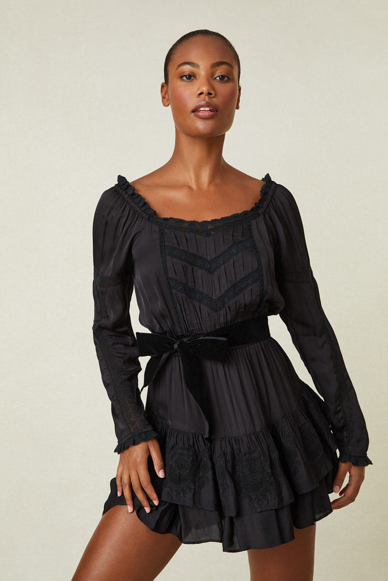 Model wearing black long sleeve mini dress with lace details and ruffled skirt. Shown here with black belt at waist.