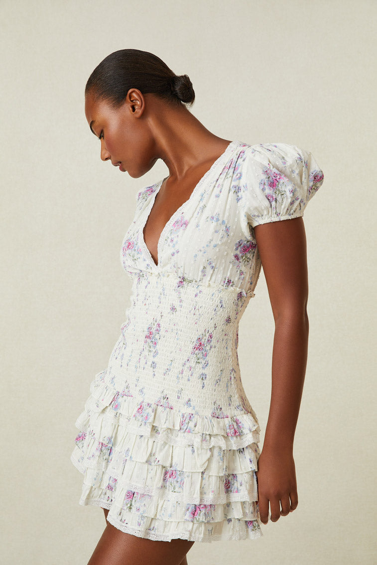 Model wearing white mini dress with pink and purple floral print. Has short puff sleeves, a smocked waistline and ruffled skirt. 