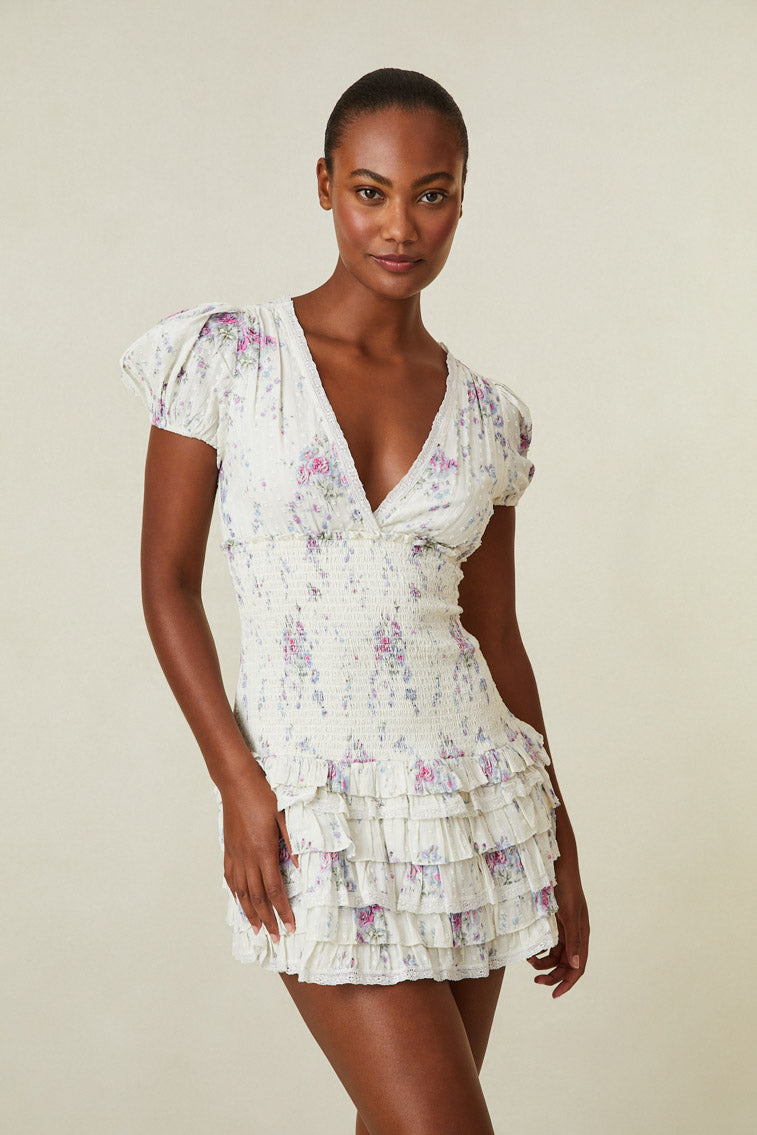 Model wearing white mini dress with pink and purple floral print. Has short puff sleeves, a smocked waistline and ruffled skirt.