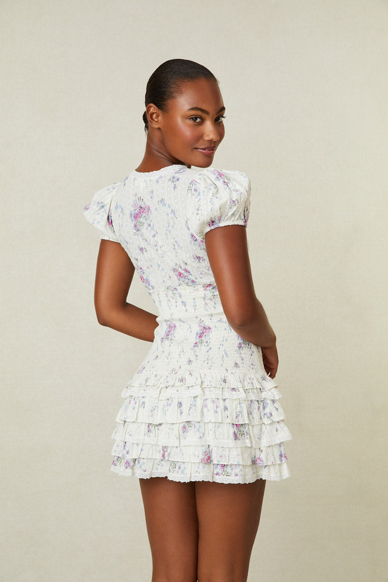 Back image of model wearing white mini dress with pink and purple floral print. Has short puff sleeves, a smocked waistline and ruffled skirt.