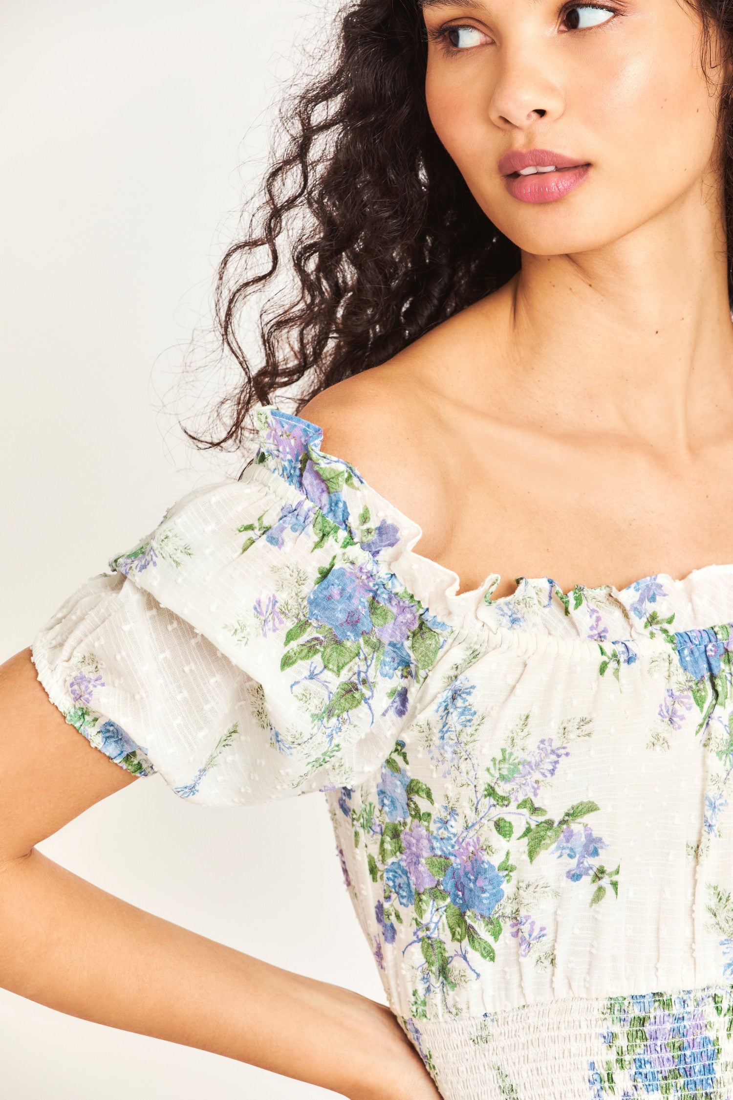 The Lai Dress in white features a blue heirloom bouquet print on a soft textured dot cotton fabric with an elasticated, ruffle-adorned neckline to be worn on or off the shoulder. It has a smocked waistband and cute puff sleeves. 