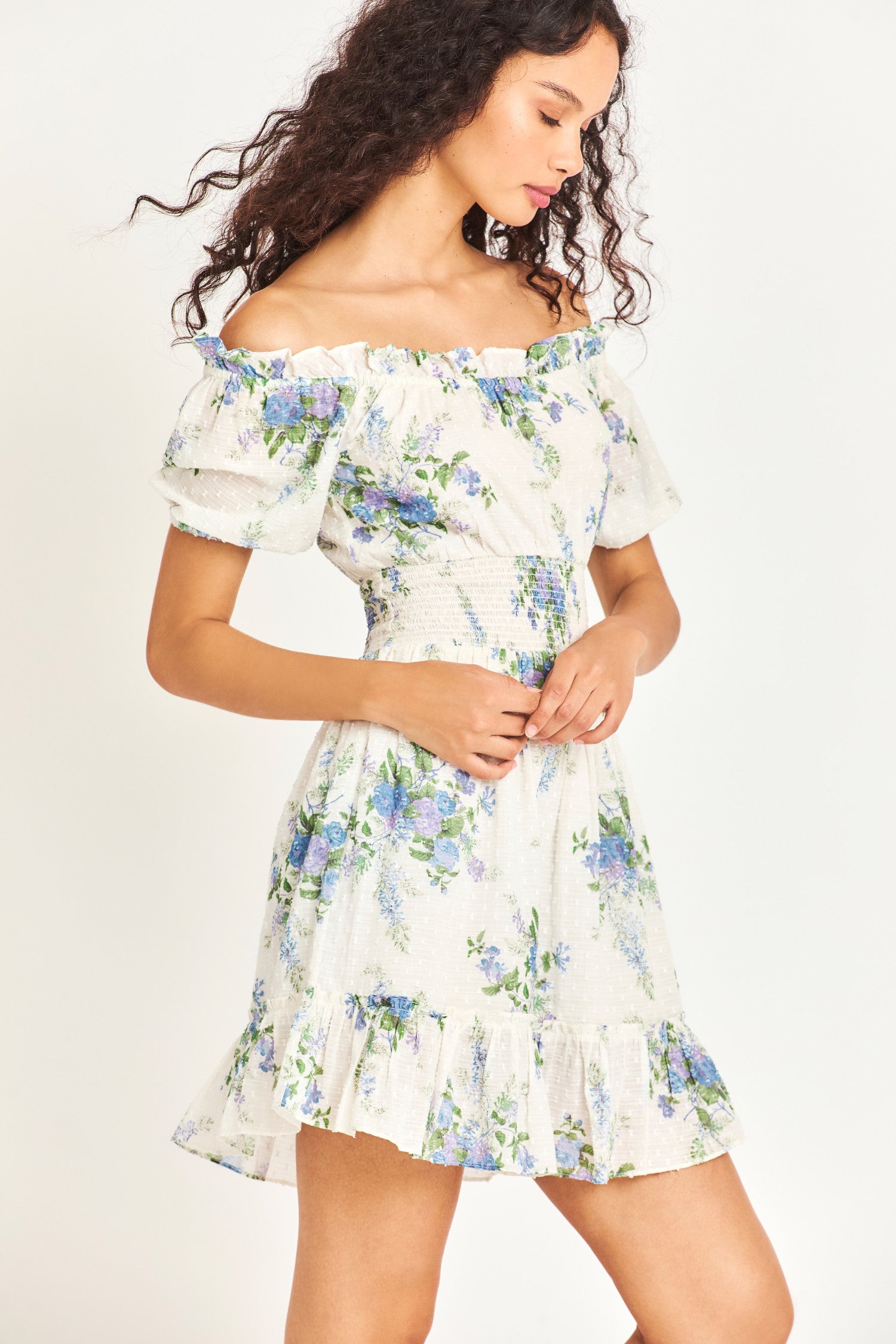 The Lai Dress in white features a blue heirloom bouquet print on a soft textured dot cotton fabric with an elasticated, ruffle-adorned neckline to be worn on or off the shoulder. It has a smocked waistband and cute puff sleeves. 