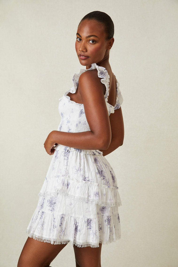 Side image of model wearing white and purple floral mini dress with smocked top and ruffled skirt