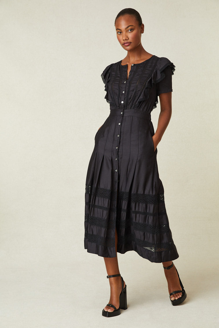 Model wearing black maxi dress with ruffle detail on shoulder, buttons down front of dress, and lace at skirt. 