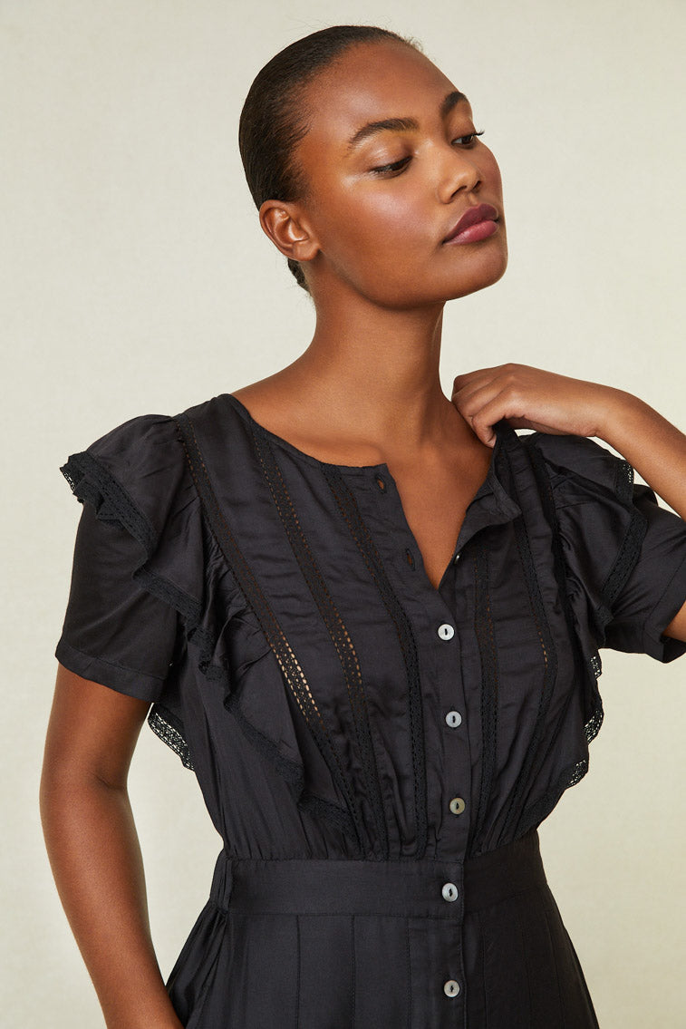 Close up image of model wearing black maxi dress with ruffle detail on shoulder, buttons down front of dress, and lace at skirt.