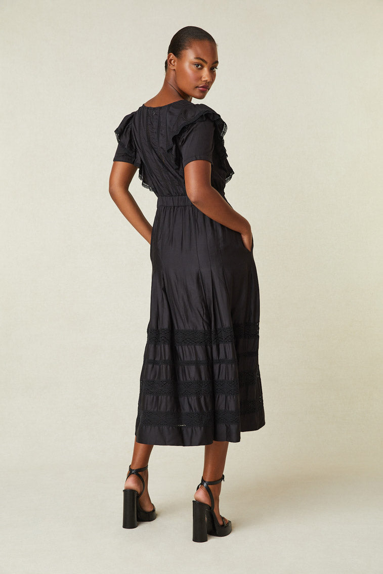 Back image of model wearing black maxi dress with ruffle detail on shoulder, buttons down front of dress, and lace at skirt.
