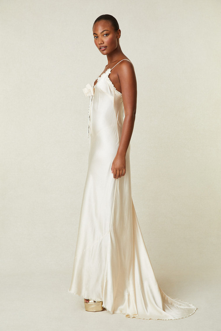 Side image of model wearing cream silk maxi slip dress with removable rosette pin and raw hem
