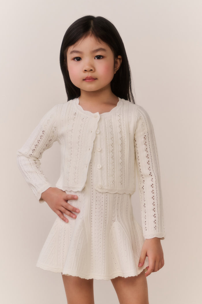 Young girl wearing a cream-colored knit cardigan and skirt outfit against a plain background