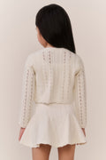 Young girl wearing a cream-colored knit cardigan and skirt outfit against a plain background