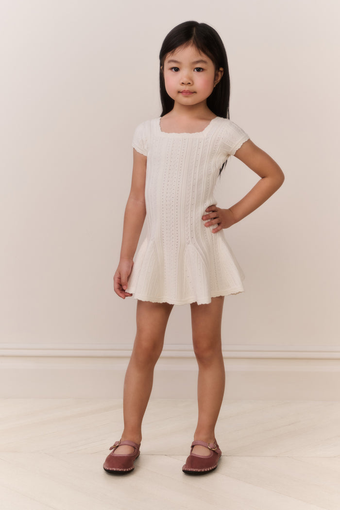 Young girl wearing a white knit dress standing against a plain background