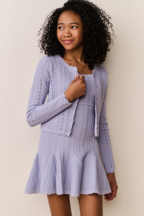 Young girl wearing a light purple dress and cardigan against a plain background