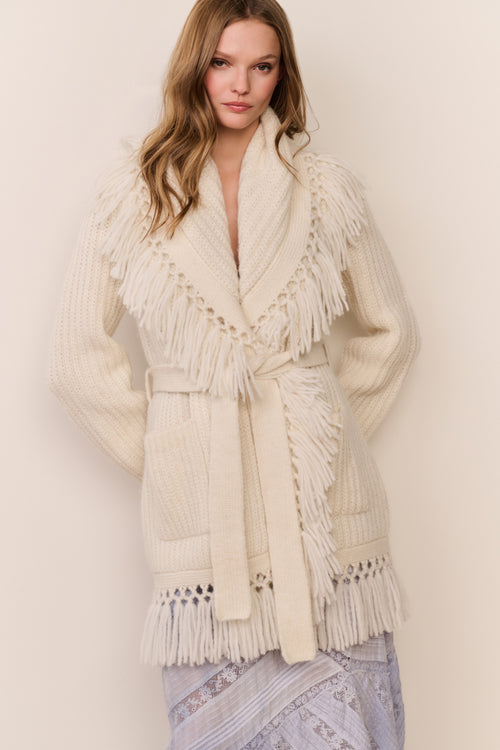 Woman wearing a cream-colored fringed cardigan against a plain background