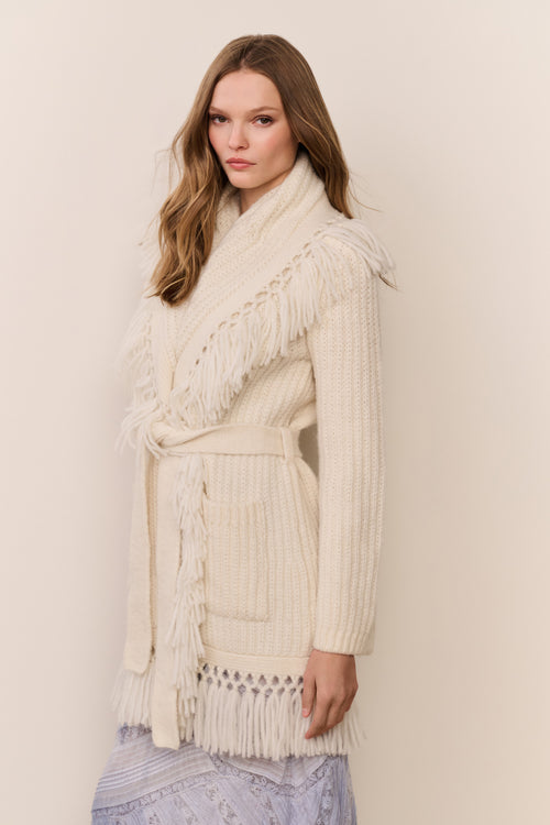 Woman wearing a cream-colored fringed sweater against a plain background