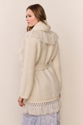 Woman wearing a cream-colored knit cardigan with fringe details on a plain background