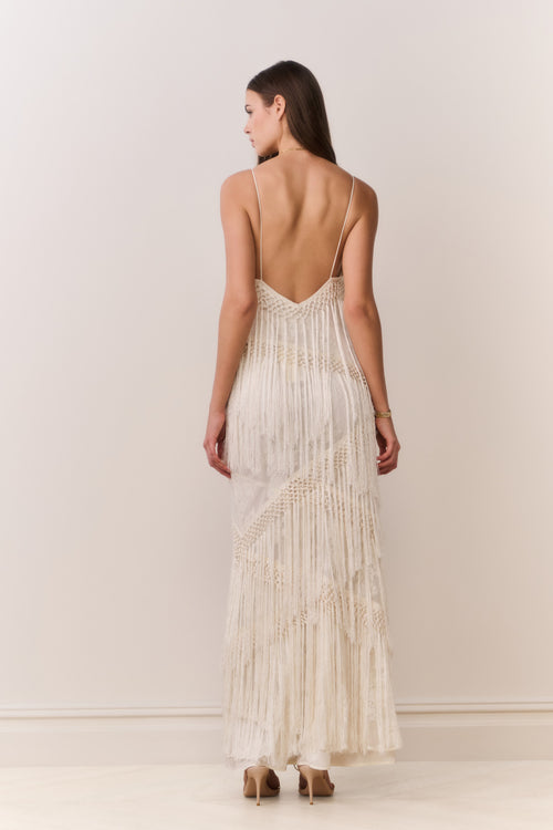 Woman wearing a long, flowing fringe dress with a backless design on a plain background