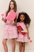 Two young girls in pink dresses holding heart-shaped items against a light background