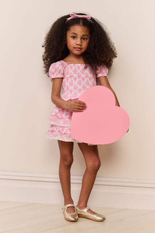 Young girl in a pink dress holding a heart-shaped object against a plain background