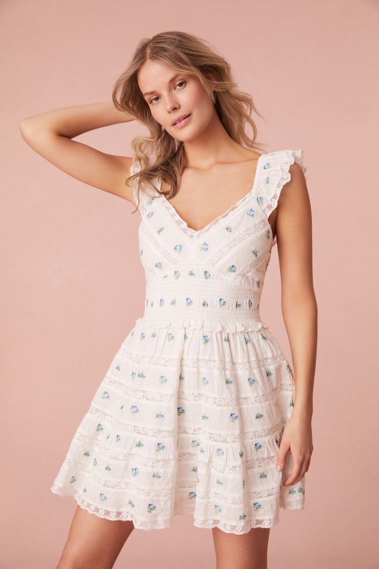 Mini dress featuring a pocono ikat floral print. The dress begins with an open v-neck with elasticated flutter sleeves before falling to a smocked waist. The bodice and skirt features lace and embroidery arrangements throughout. This piece is complete with pockets.