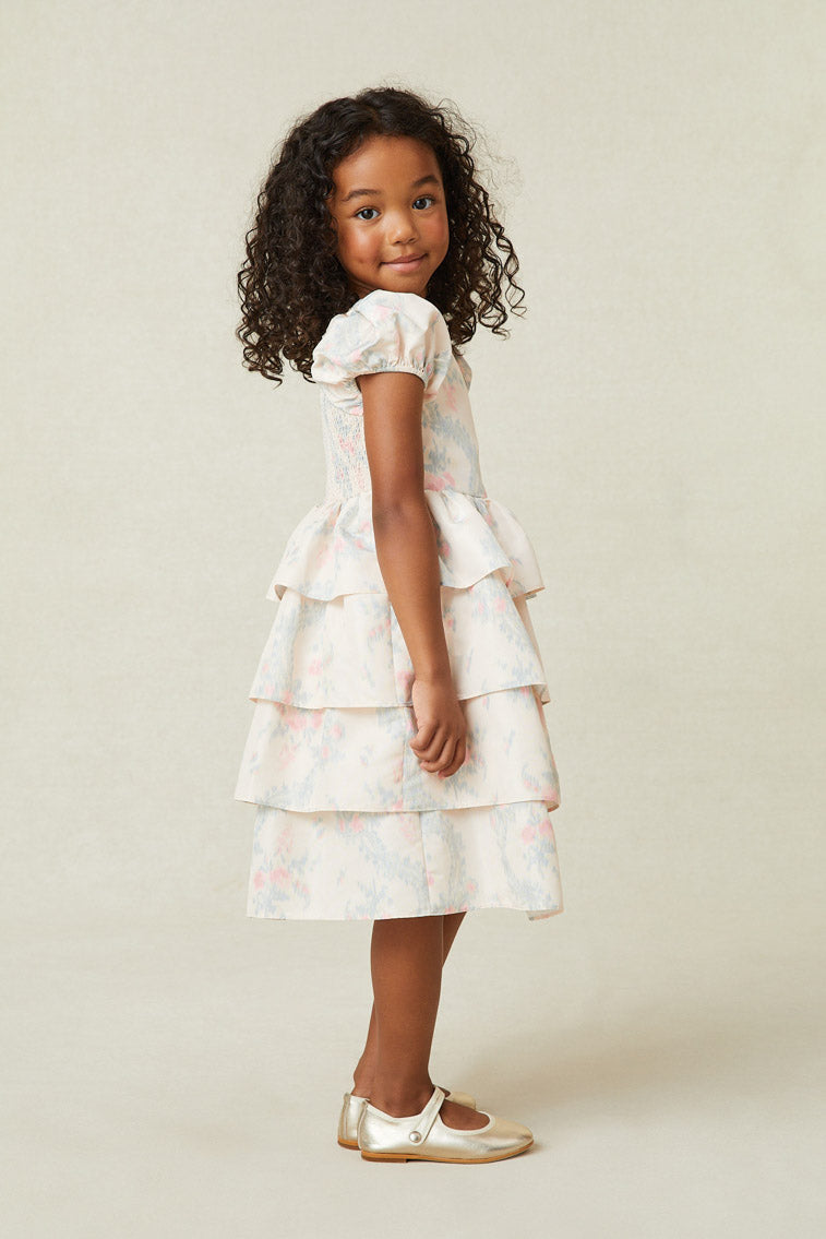 Side image of model wearing girl's light pink midi dress with blue and pink floral print. Has puff sleeves and a tiered ruffle skirt.