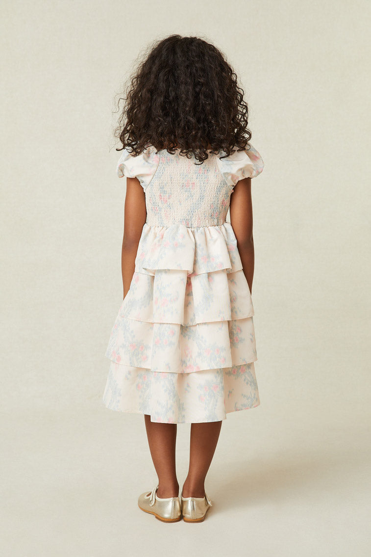 Back image of model wearing girl's light pink midi dress with blue and pink floral print. Has puff sleeves and a tiered ruffle skirt.