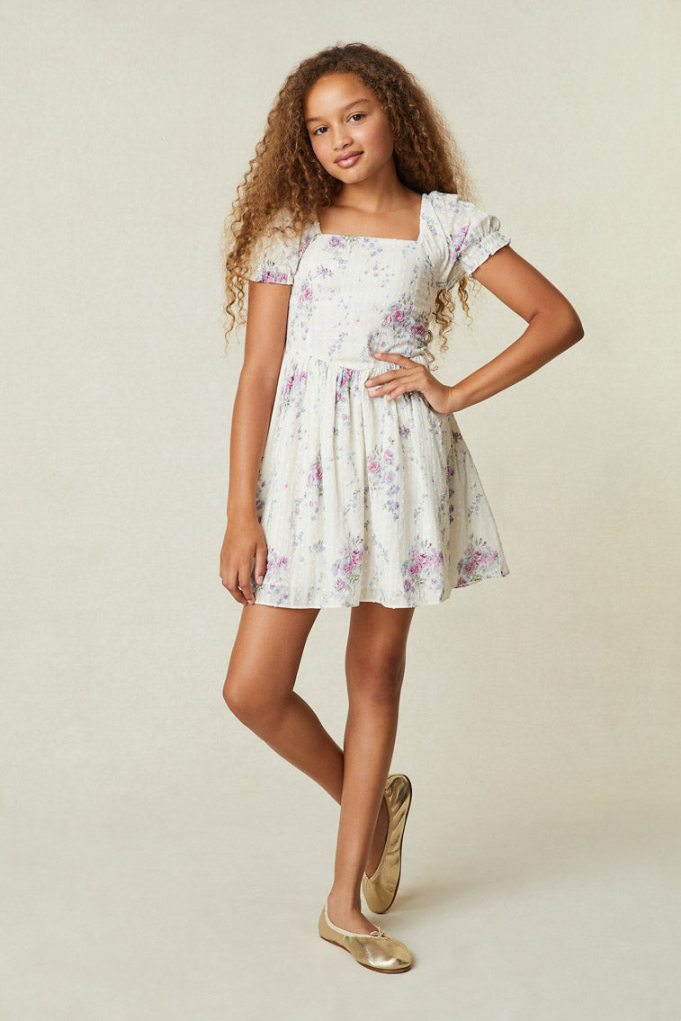Model wearing girl's white mini dress with purple and pink floral print. 