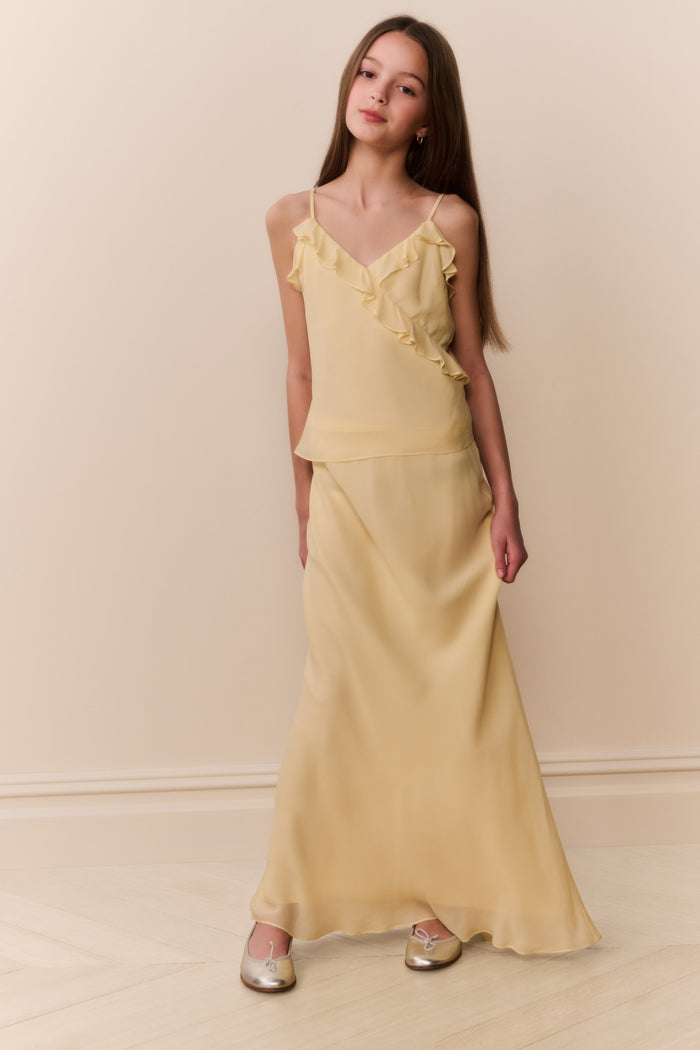 Girl wearing a light yellow sleeveless ruffle detail top and matching long skirt against a beige background
