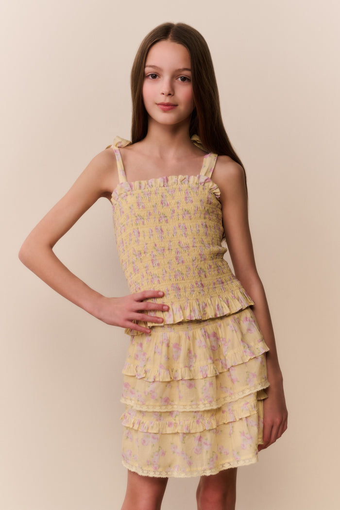 Tween girl wearing a yellow floral ruffle top and matching skirt with tiered layers on a beige background.