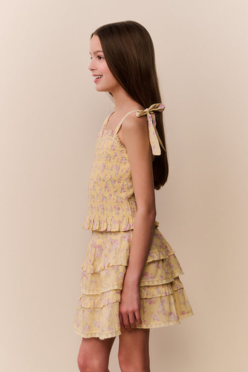 Tween girl wearing a yellow floral ruffle top and matching skirt with tiered layers on a beige background.